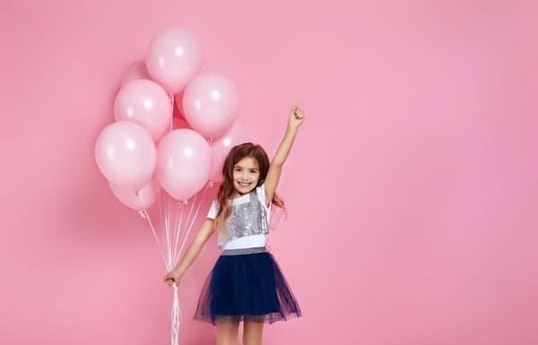 A happy and fashionable young girl is posing with pastel pink birthday balloons on a pink background.