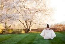 A teenage girl, dressed in a pink ballroom gown for her quinceañera, is walking away while posing next to pink Jasmine trees.