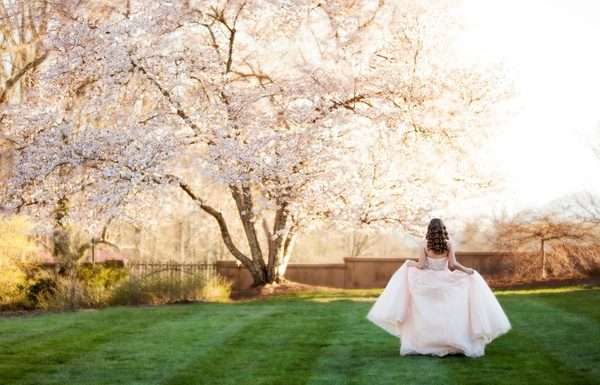 A teenage girl, dressed in a pink ballroom gown for her quinceañera, is walking away while posing next to pink Jasmine trees.