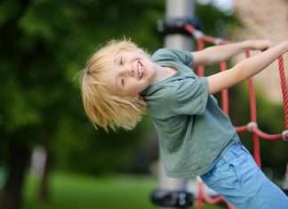 The Best Hard-Weather Clothes for High-Energy Kids A smiling child, dressed in a green t-shirt and jeans, playfully hanging off of a rope section on a playground.