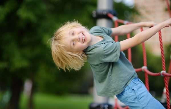 A smiling child, dressed in a green t-shirt and jeans, playfully hanging off of a rope section on a playground.