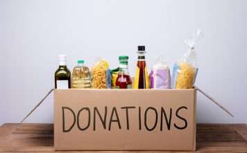 A cardboard box with "DONATIONS" written in black marker on the side. The box is filled with pasta, olive oil, and rice.