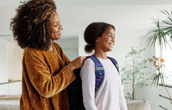 Calming Rituals To Start Every Busy Morning Right A mother and her daughter are smiling and talking as the mother helps her child put her backpack on for school.