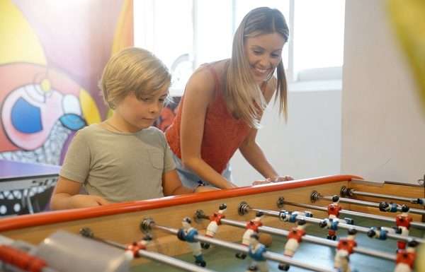 A woman and a young boy smile while playing foosball together at a table in a bright indoor recreation area.