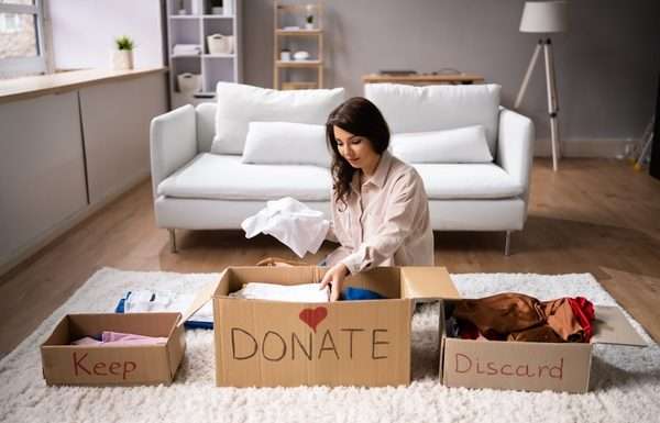 A woman sits on the floor in front of a white couch and sorts clothing into boxes labeled, "Keep," "DONATE," and "Discard."
