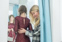 Mother helping her daughter try on a sparkling dress in a fitting room while checking the look in a mirror.