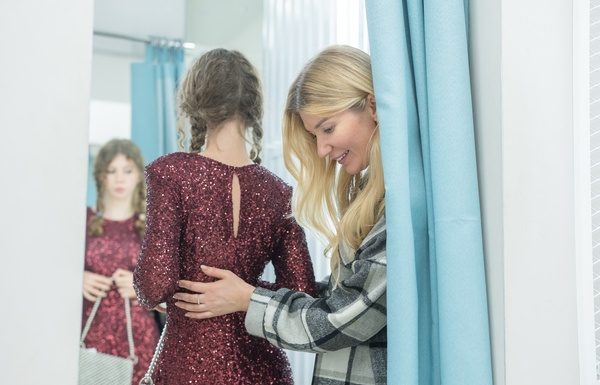 Mother helping her daughter try on a sparkling dress in a fitting room while checking the look in a mirror.