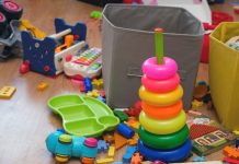 A playroom is cluttered with kids' toys, including a ring stacker, puzzle pieces, a toy xylophone, a book, and two bins.
