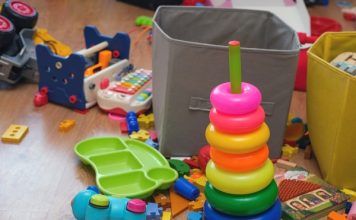 A playroom is cluttered with kids' toys, including a ring stacker, puzzle pieces, a toy xylophone, a book, and two bins.