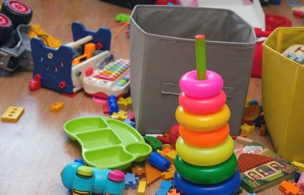 A playroom is cluttered with kids' toys, including a ring stacker, puzzle pieces, a toy xylophone, a book, and two bins.