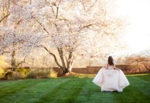 A teen girl in a pale formal dress walking across a grassy lawn toward blossoming trees in warm sunlight.