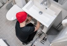 A man in a beanie using tools to adjust something below a bathroom sink. The floor is littered with other tools.