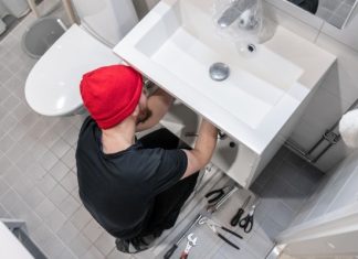 Everything You Need To Do Bathroom Remodeling A man in a beanie using tools to adjust something below a bathroom sink. The floor is littered with other tools.