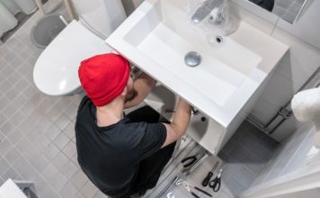 A man in a beanie using tools to adjust something below a bathroom sink. The floor is littered with other tools.