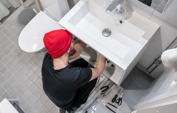 A man in a beanie using tools to adjust something below a bathroom sink. The floor is littered with other tools.