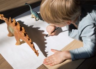 A child drawing on white paper with a pencil beside toy dinosaur figures, casting shadows on the surface.