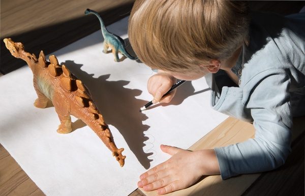 Fun Ways to Spark Your Kid’s Love of Dinosaurs A child drawing on white paper with a pencil beside toy dinosaur figures, casting shadows on the surface.