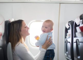 How To Survive Travel Abroad with Young Kids A young mother holding her baby in front of her as she sits on a plane and both smile at one another.