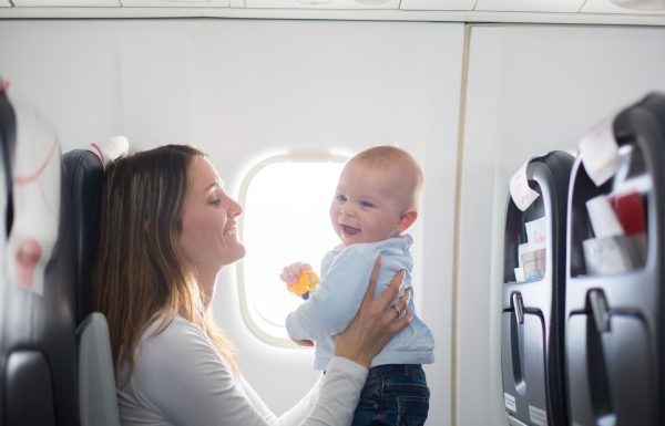A young mother holding her baby in front of her as she sits on a plane and both smile at one another.