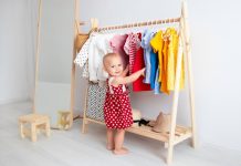 A smiling toddler wearing a red polka dot dress while standing beside a wooden rack of colorful clothing.