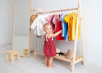 A smiling toddler wearing a red polka dot dress while standing beside a wooden rack of colorful clothing.