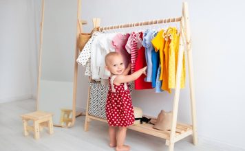 A smiling toddler wearing a red polka dot dress while standing beside a wooden rack of colorful clothing.