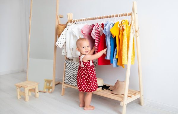 5 Fun Pieces to Add to Your Toddler’s Wardrobe A smiling toddler wearing a red polka dot dress while standing beside a wooden rack of colorful clothing.