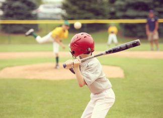 How To Help Your Kid Improve as a Baseball/Softball Hitter A view from behind of a young baseball player with a red helmet and black bat preparing for an incoming pitch.