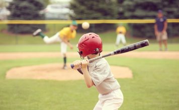 A view from behind of a young baseball player with a red helmet and black bat preparing for an incoming pitch.