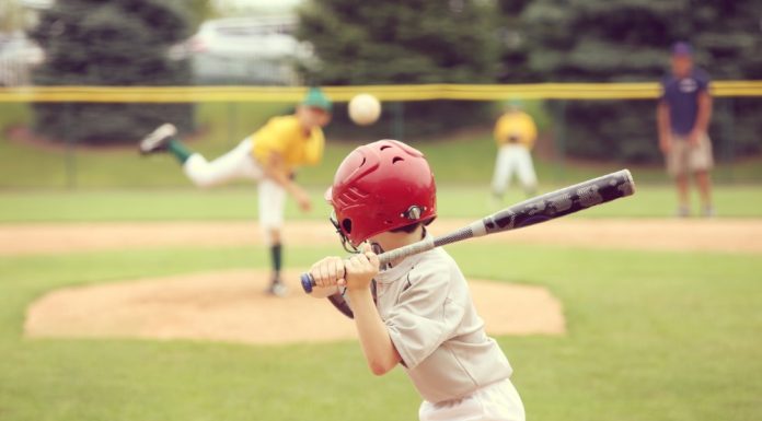 A view from behind of a young baseball player with a red helmet and black bat preparing for an incoming pitch.
