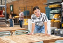 Why Winter Is the Ideal Time To Buy Patio Furniture A woman in a striped shirt and jeans leans to touch a wooden outdoor table display in a furniture store.