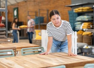 A woman in a striped shirt and jeans leans to touch a wooden outdoor table display in a furniture store.