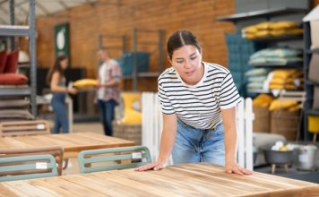 A woman in a striped shirt and jeans leans to touch a wooden outdoor table display in a furniture store.