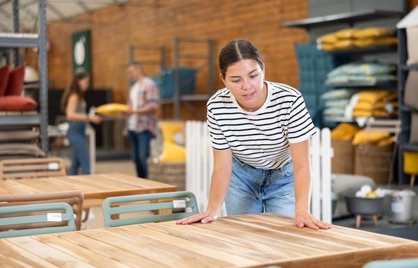 A woman in a striped shirt and jeans leans to touch a wooden outdoor table display in a furniture store.