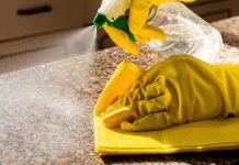 How To Protect Stone Countertops From Daily Wear A close-up of a hands with yellow cleaning gloves using a spray bottle and yellow cloth to clean a granite countertop.