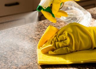 How To Protect Stone Countertops From Daily Wear A close-up of a hands with yellow cleaning gloves using a spray bottle and yellow cloth to clean a granite countertop.