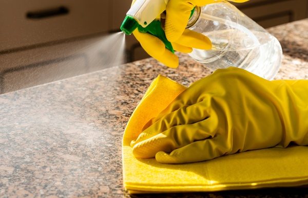 A close-up of a hands with yellow cleaning gloves using a spray bottle and yellow cloth to clean a granite countertop.